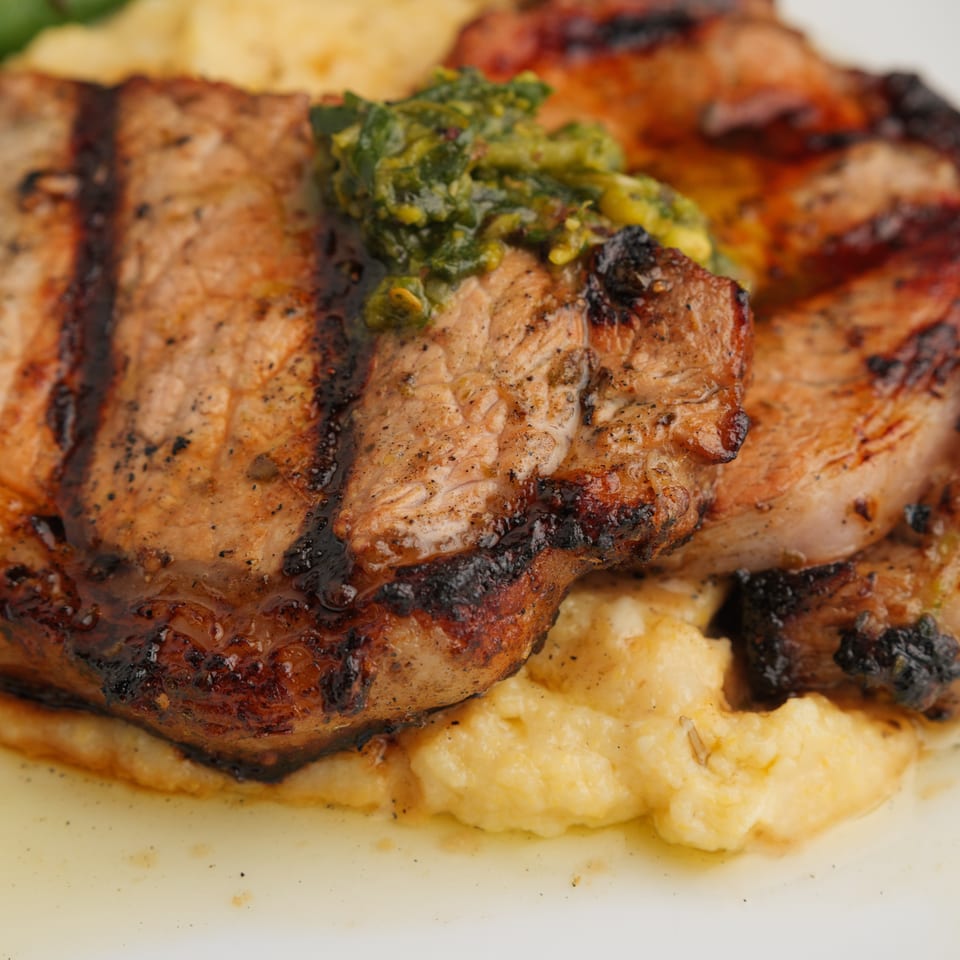 Grilled pork chops with green herb sauce on a bed of polenta, with green beans in the background.