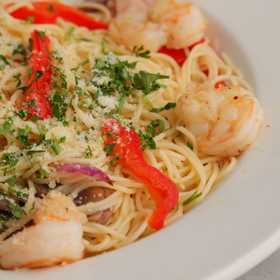 Close-up of shrimp scampi with spaghetti, red bell peppers, mushrooms, and parsley in a white bowl.