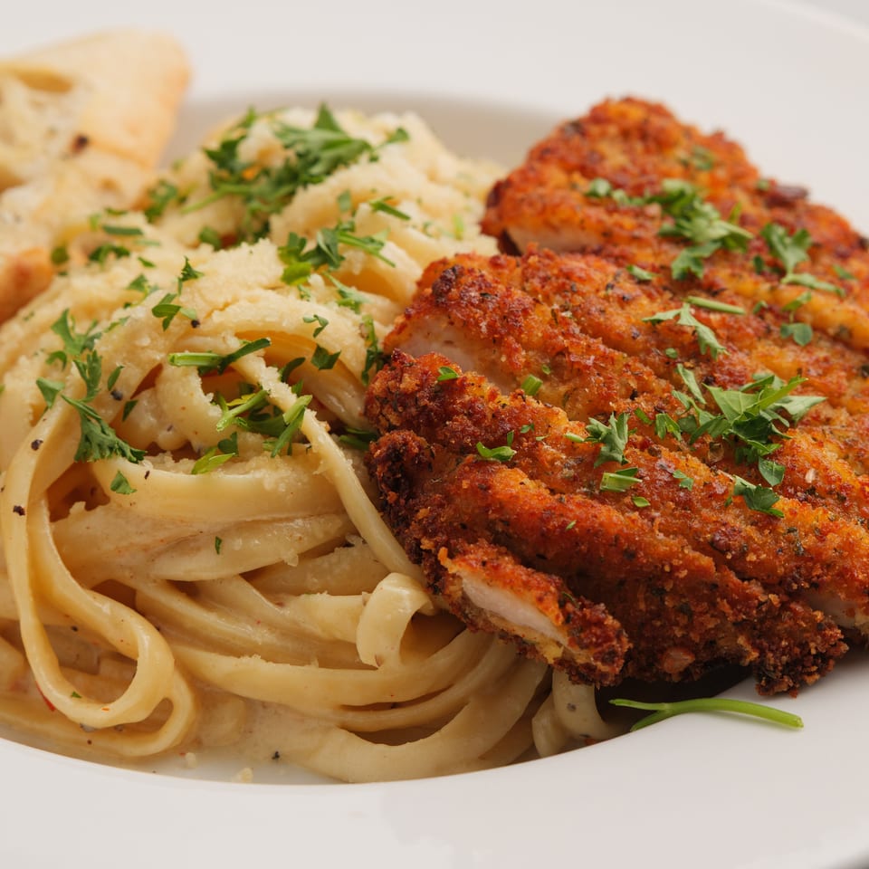 Crispy breaded chicken cutlets with creamy fettuccine alfredo, garnished with parsley and garlic bread.